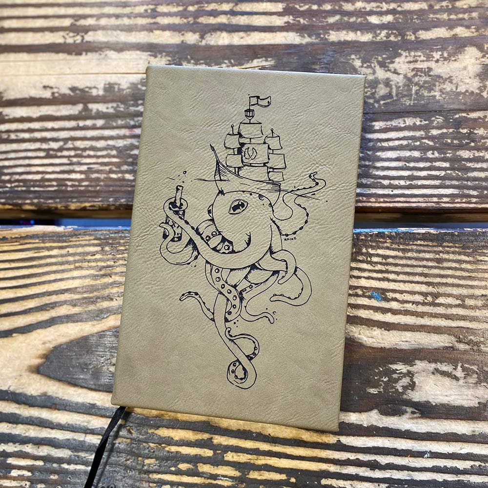 8% salty faux leather journal with octopus, ship, and bottle lying on wooden table.
