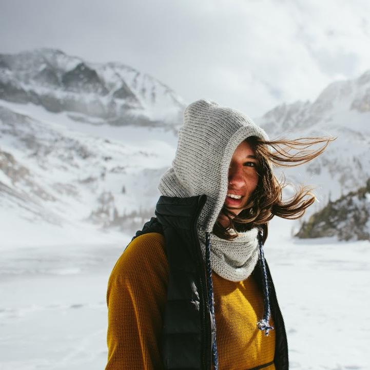 Woman in yellow knit sweater and vest wears our Verse Hooded Cowl in front of snowy winter landscape