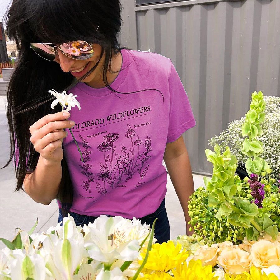 woman smelling flowers in a purple tee printed with a colorado wildflowers design