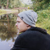man wearing light grey ahab knitted beanie and black hoodie sitting by river