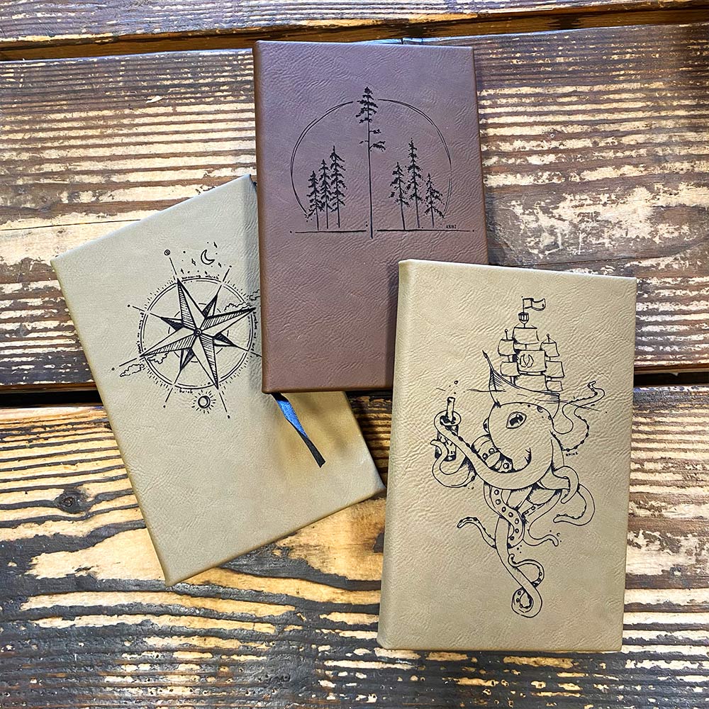Find your path compass faux leather journal, speak for the trees dark brown faux leather journal, and 8% salty faux leather journal lying on wooden table.